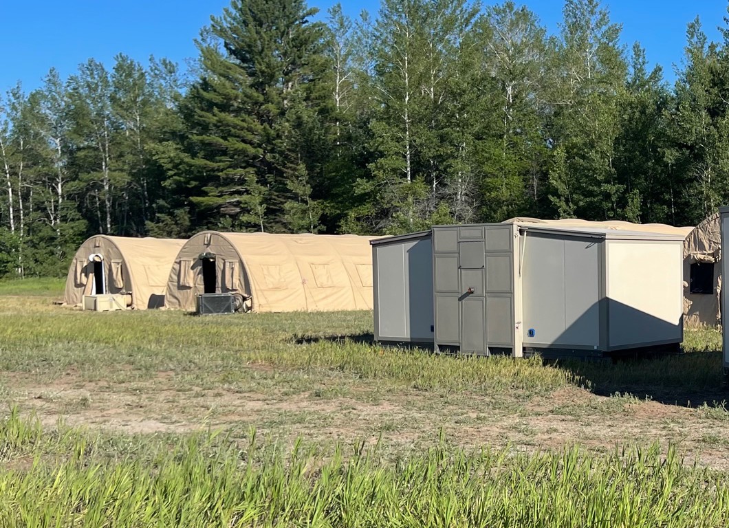 Actual white ACE shelter deployed on grass with beige tents behind it