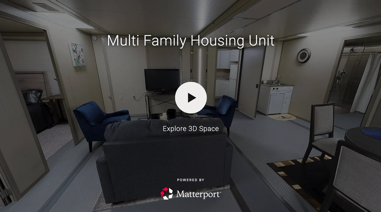 Interior view of a multi-family housing unit with a living room, television, blue chairs, a dining table with chairs, a kitchenette, and a bathroom with a shower.