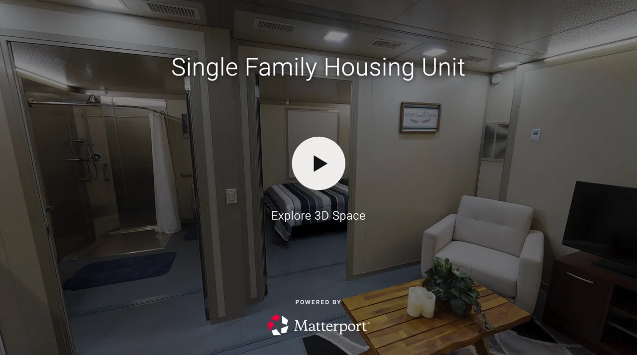 Interior of a single-family housing unit with a small living space, a glimpse of a bedroom with a bed, a bathroom with a shower, and some furniture including a white armchair and a wooden coffee table with candles and a plant.